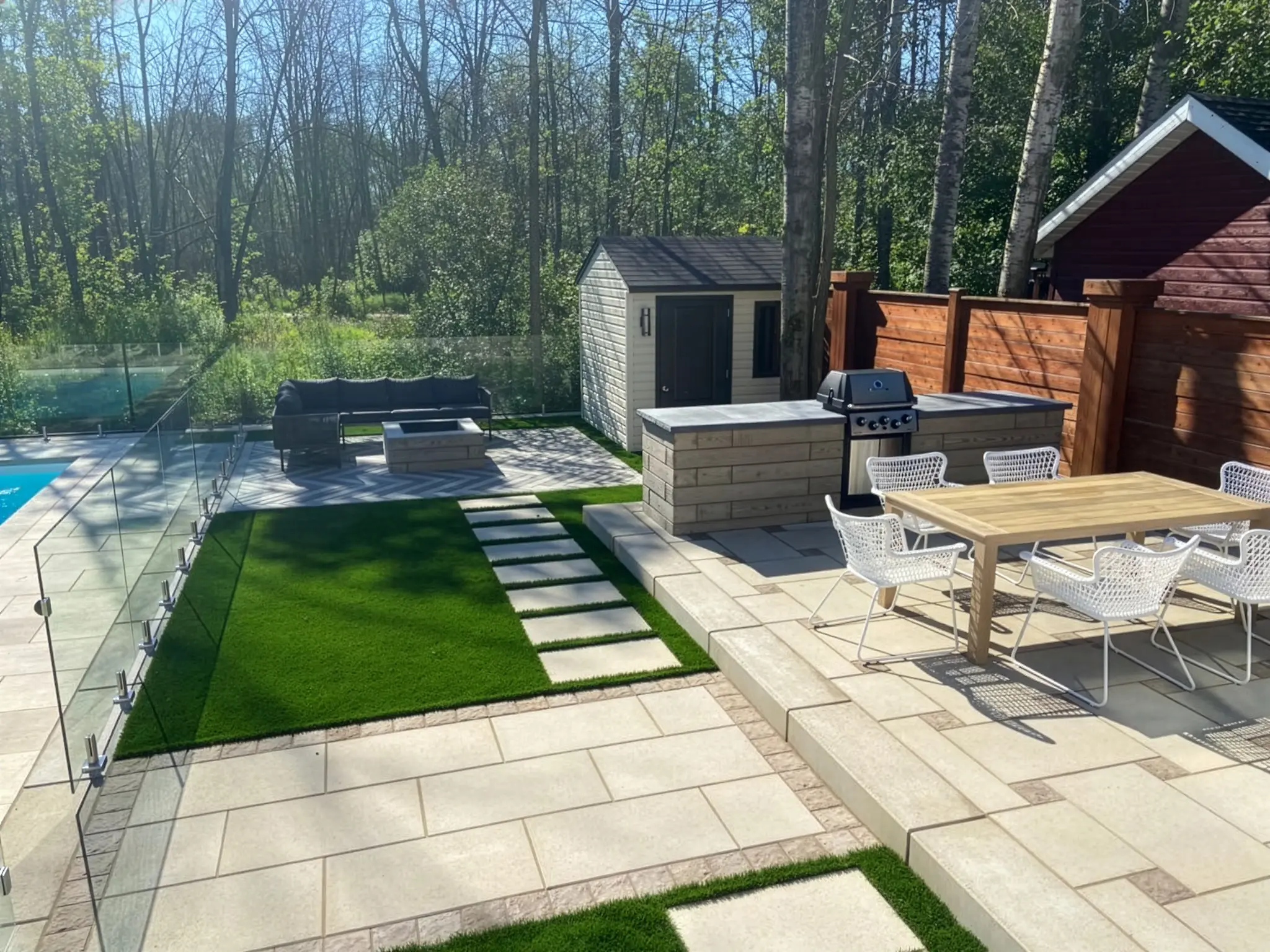 Aménagement complet autour de piscine avec pavé uni, gazon artificiel, cuisine extérieure et clôture de verre